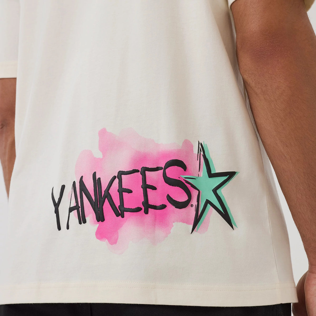 New Era New York Yankees MLB Street Art Graphic Light Beige T-Shirt, back view showing Yankees lettering with a black finish over a pink background and green star graphic near the bottom hem, styled casually with dark trousers.