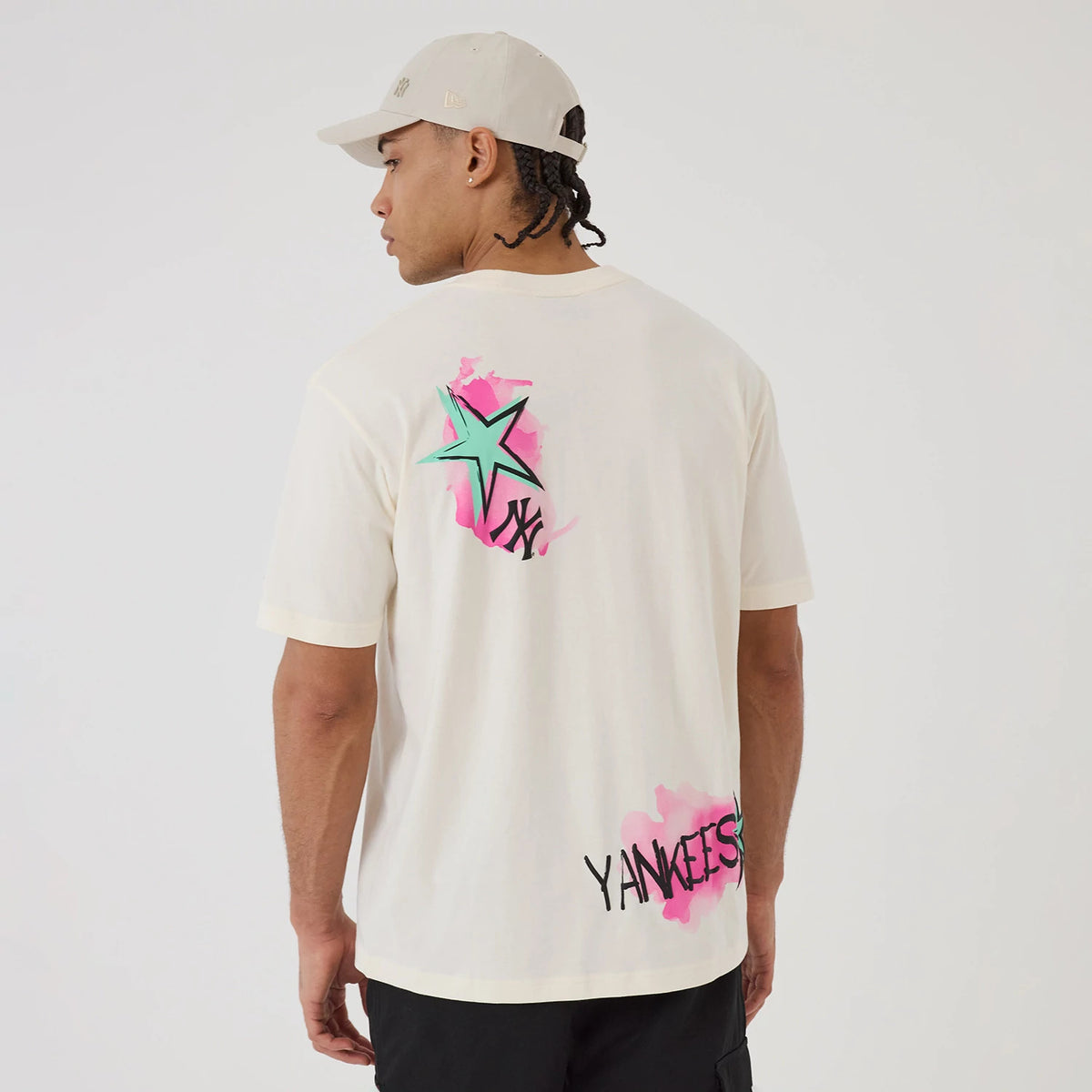 New Era New York Yankees MLB Street Art Graphic Light Beige T-Shirt, back view on a model with a cap, showing a green and pink star graphic, NY lettering, and Yankees text in black against a pink splash on the lower side.