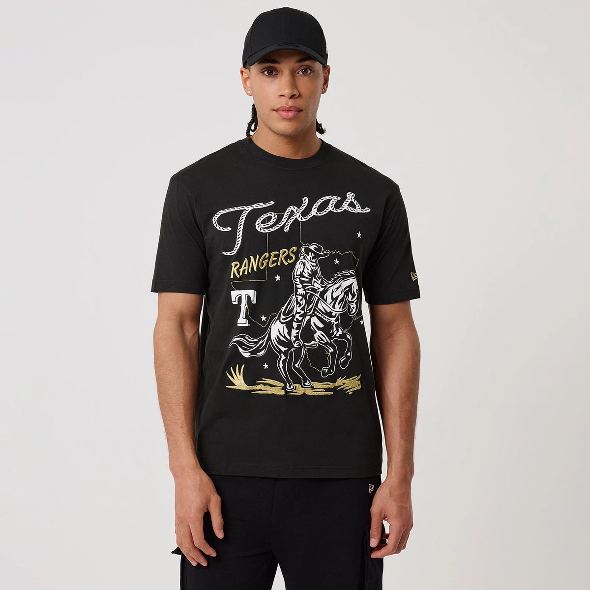 New Era Texas Rangers MLB Cowboy Graphic Black T-Shirt, front view on a model with a plain black cap, features Texas lettering, golden Rangers text, a cowboy on horseback graphic, and a printed New Era logo on the left sleeve.
