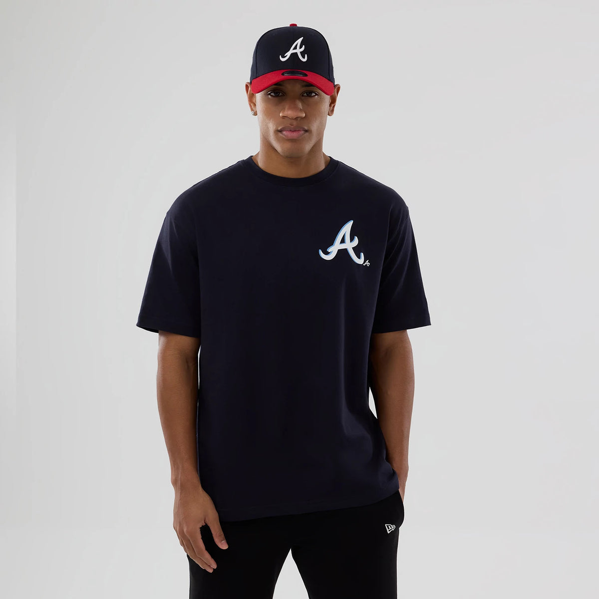 New Era Atlanta Braves MLB Graphic Navy T-Shirt, front view on-model showing the large white embroidered A logo on the left chest, with the navy T-shirt styled alongside a coordinating navy and red cap featuring a matching logo.