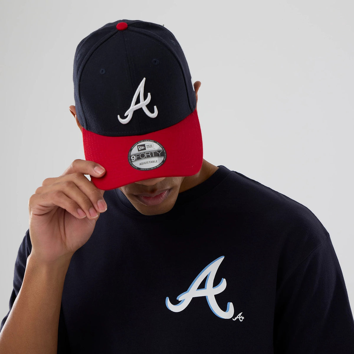 New Era Atlanta Braves MLB Graphic Navy T-Shirt, on-model view featuring a large white printed Braves logo on the front left side, styled with a navy and red cap showing an embroidered white A on the front panel.