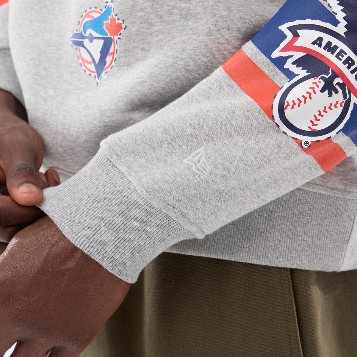 New Era Toronto Blue Jays MLB Sport Classic Grey Crew Neck Sweater, close-up showing the team logo printed on the chest, New Era flag logo embroidered on the ribbed cuff, and American League patch on the sleeve.