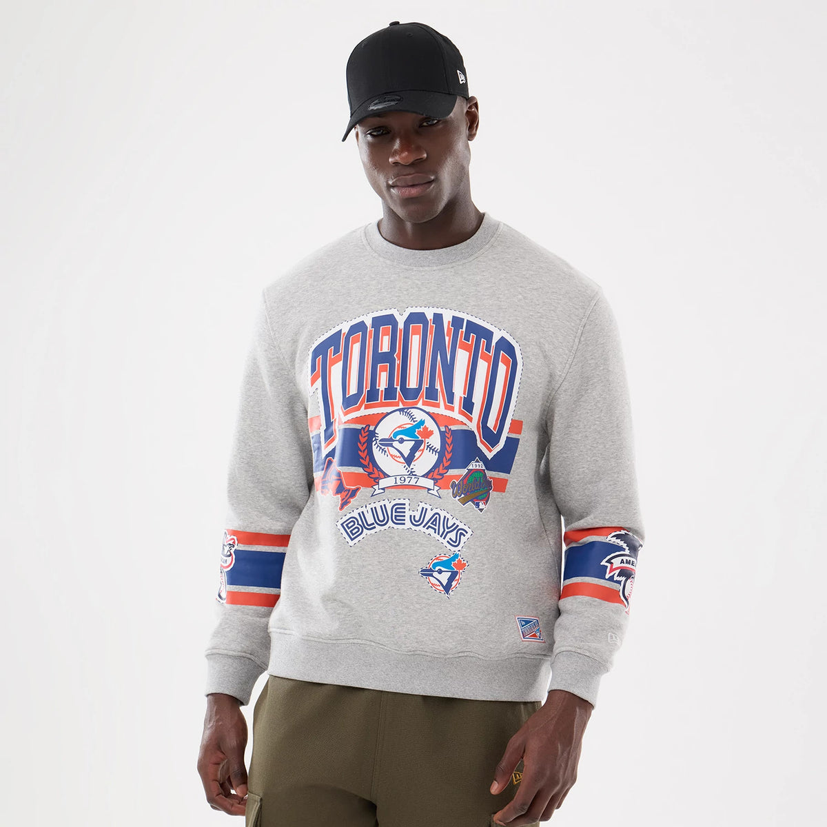 New Era Toronto Blue Jays MLB Sport Classic Grey Crew Neck Sweater, front view on a model with a black cap, featuring blue and orange Toronto lettering and Blue Jays logos on the chest and sleeves.