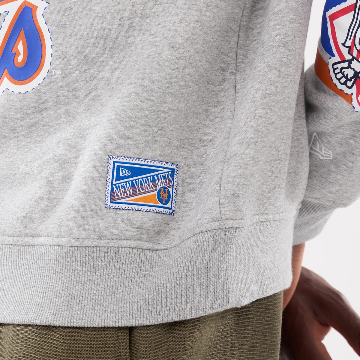 New Era New York Mets MLB Sport Classic Grey Crew Neck Sweater, side view showing an embroidered New York Mets patch near the ribbed hem and a stylised Mets graphic on the sleeve, with a small New Era flag logo.