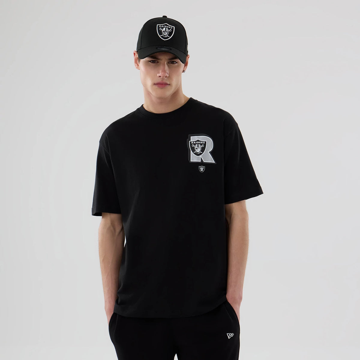 New Era Las Vegas Raiders NFL Graphic Black T-Shirt, front view on a model with a matching cap, featuring the large printed Raiders logo with a R on the left side of the chest and a smaller embroidered New Era logo near the hem.