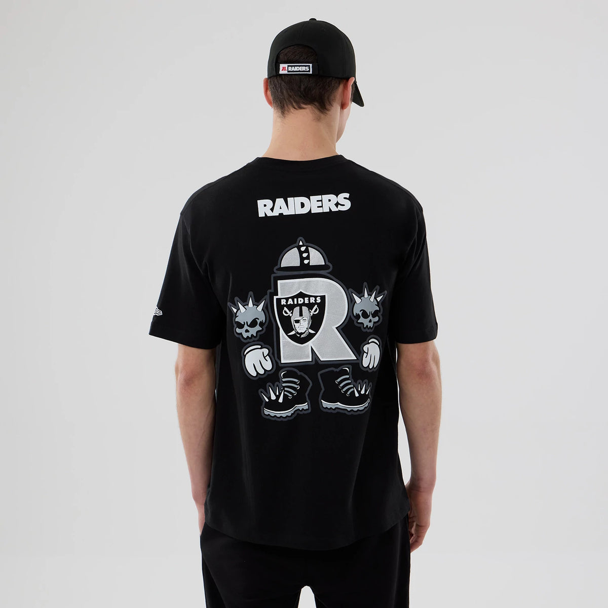 New Era Las Vegas Raiders NFL Graphic Black T-Shirt, back view on a model wearing a black cap, featuring white Raiders lettering above a large graphic design with a helmet and spiked gloves, alongside skull illustrations.
