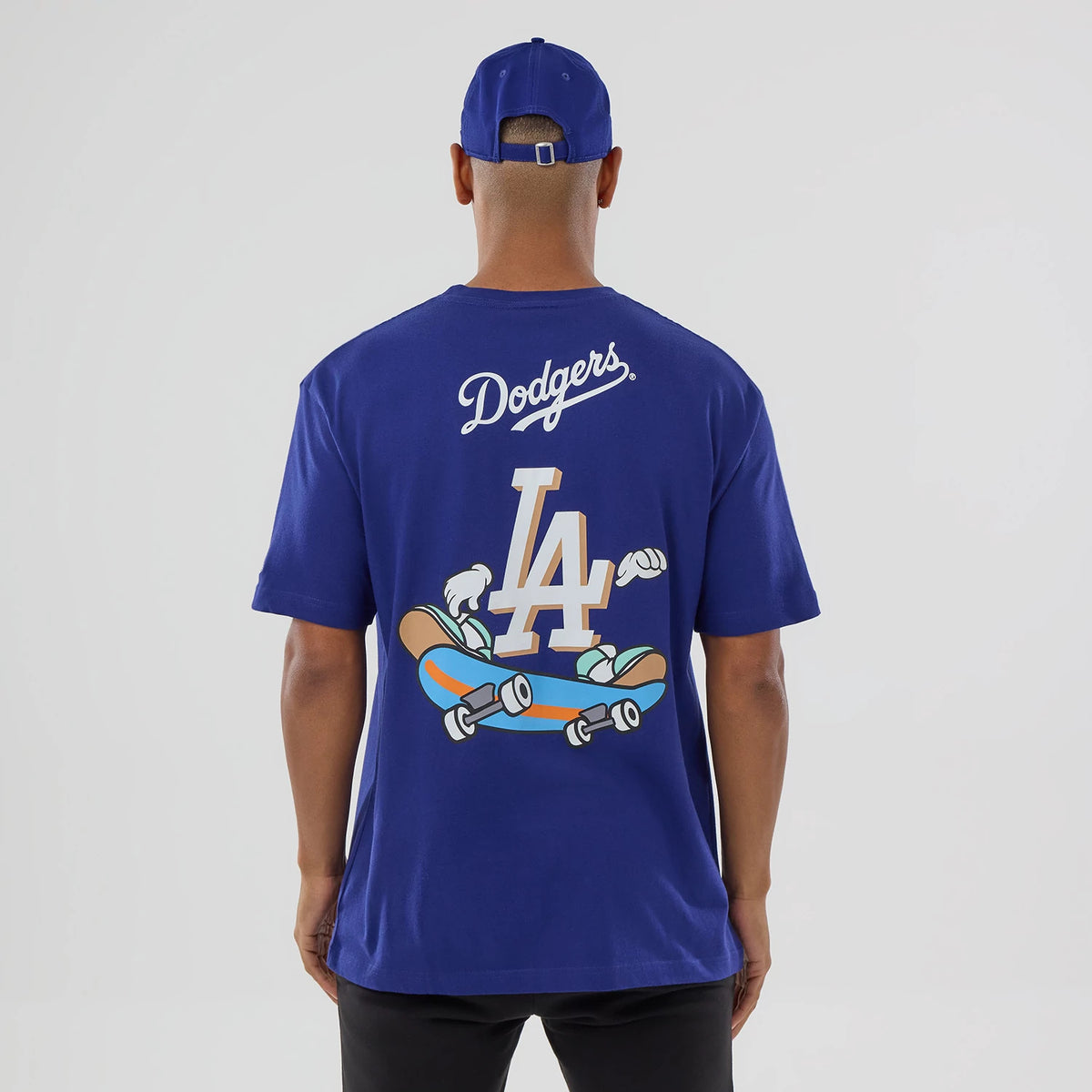 New Era LA Dodgers MLB Graphic Dark Blue T-Shirt, back view on-model showing a large printed LA logo with cartoon skateboards and gloved hands, Dodgers script logo across the shoulders, and matching cap styled with the shirt.