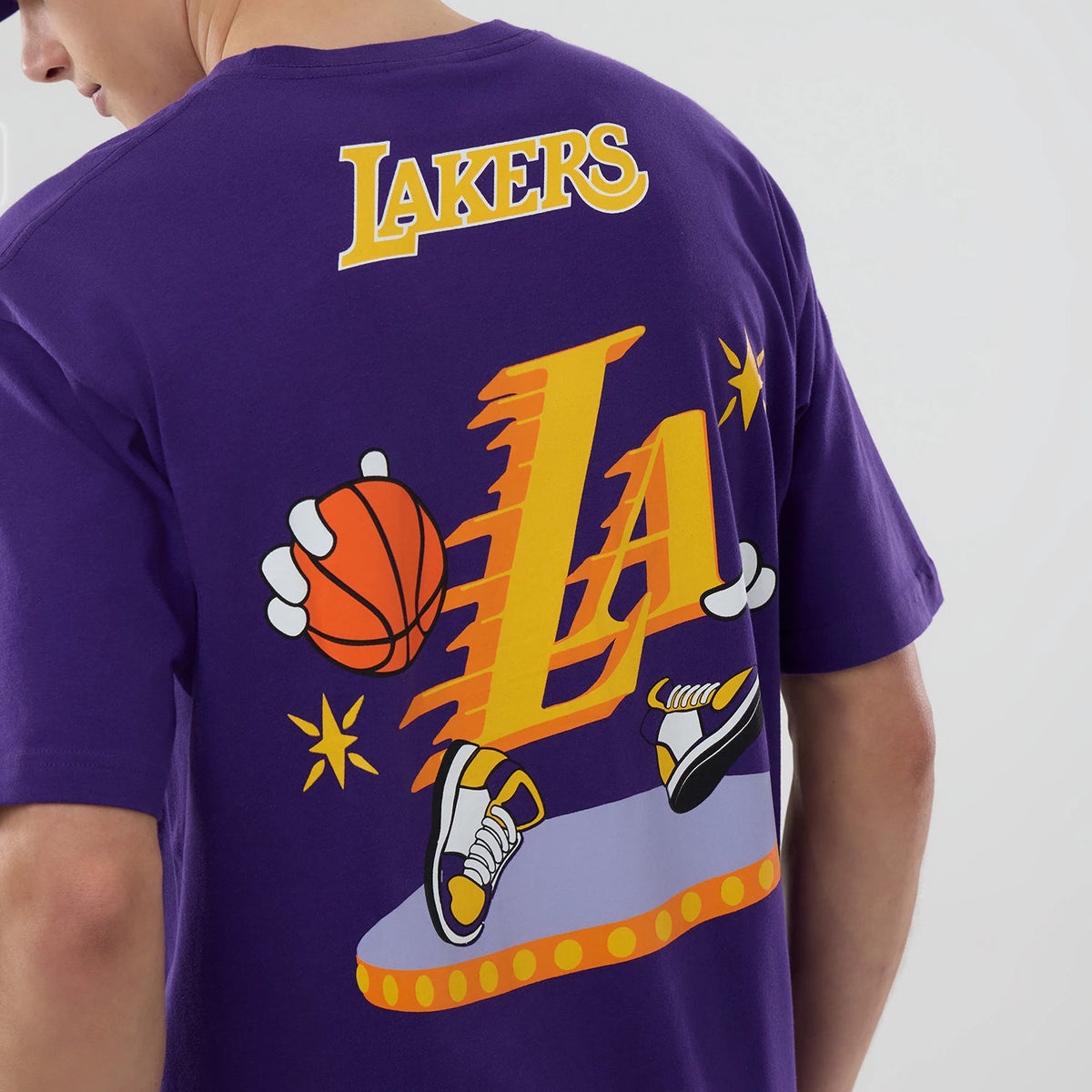 New Era LA Lakers NBA Graphic Purple T-Shirt, back view on a model showing Lakers lettering above a large graphic of shoes, basketball, and stars with an orange and yellow LA icon in the centre.