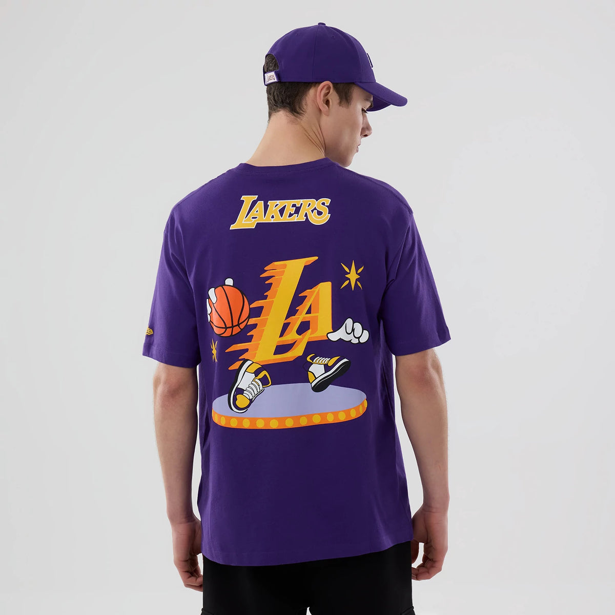 New Era LA Lakers NBA Graphic Purple T-Shirt, back view on a model wearing a purple cap and black shorts, featuring a graphic of a basketball and sneakers with large gold LA lettering and Lakers text above.