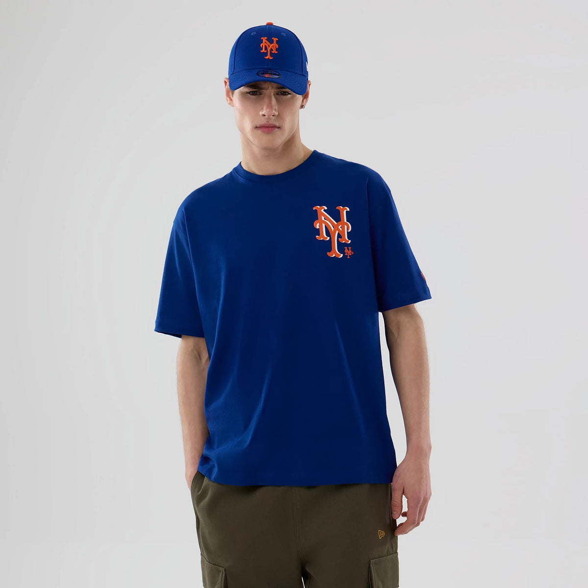 New Era New York Mets MLB Graphic Blue T-Shirt, front view on a model featuring a large embroidered orange and white NY logo on the left chest, paired with a matching blue cap displaying the same logo, against a plain background.