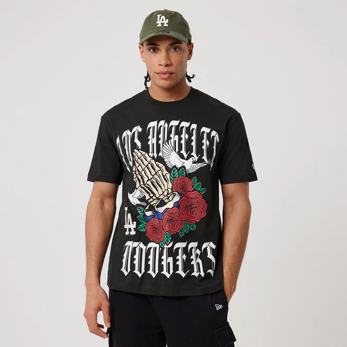 New Era LA Dodgers MLB Script Graphic Black T-Shirt, front view on-model showing the intricate graphic of praying hands, roses, and doves with Los Angeles Dodgers lettering, and a small embroidered New Era logo on the sleeve.