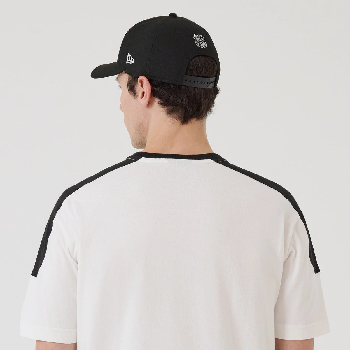 New Era LA Kings NHL Sport Classic White T-Shirt, back view showing the plain white back with black shoulder panels, worn with a black cap featuring an embroidered New Era flag logo on the side and an NHL logo on the back.