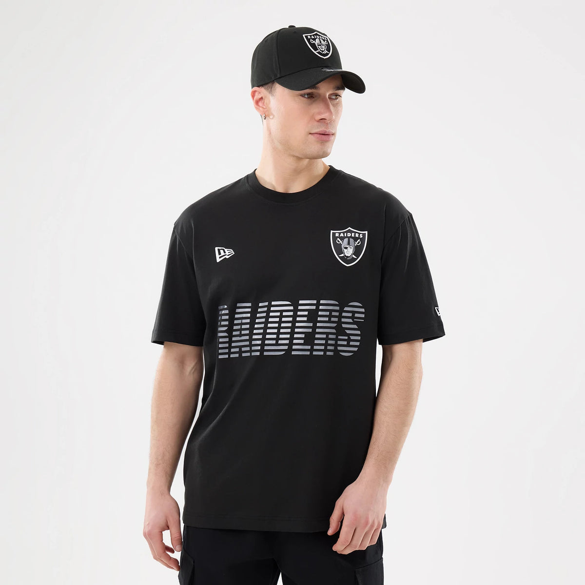 New Era Las Vegas Raiders NFL Match Black T-Shirt, front view on a model wearing a matching cap, showing a raised Raiders shield logo on the chest, a printed Raiders wordmark below, and an embroidered New Era logo on the right.