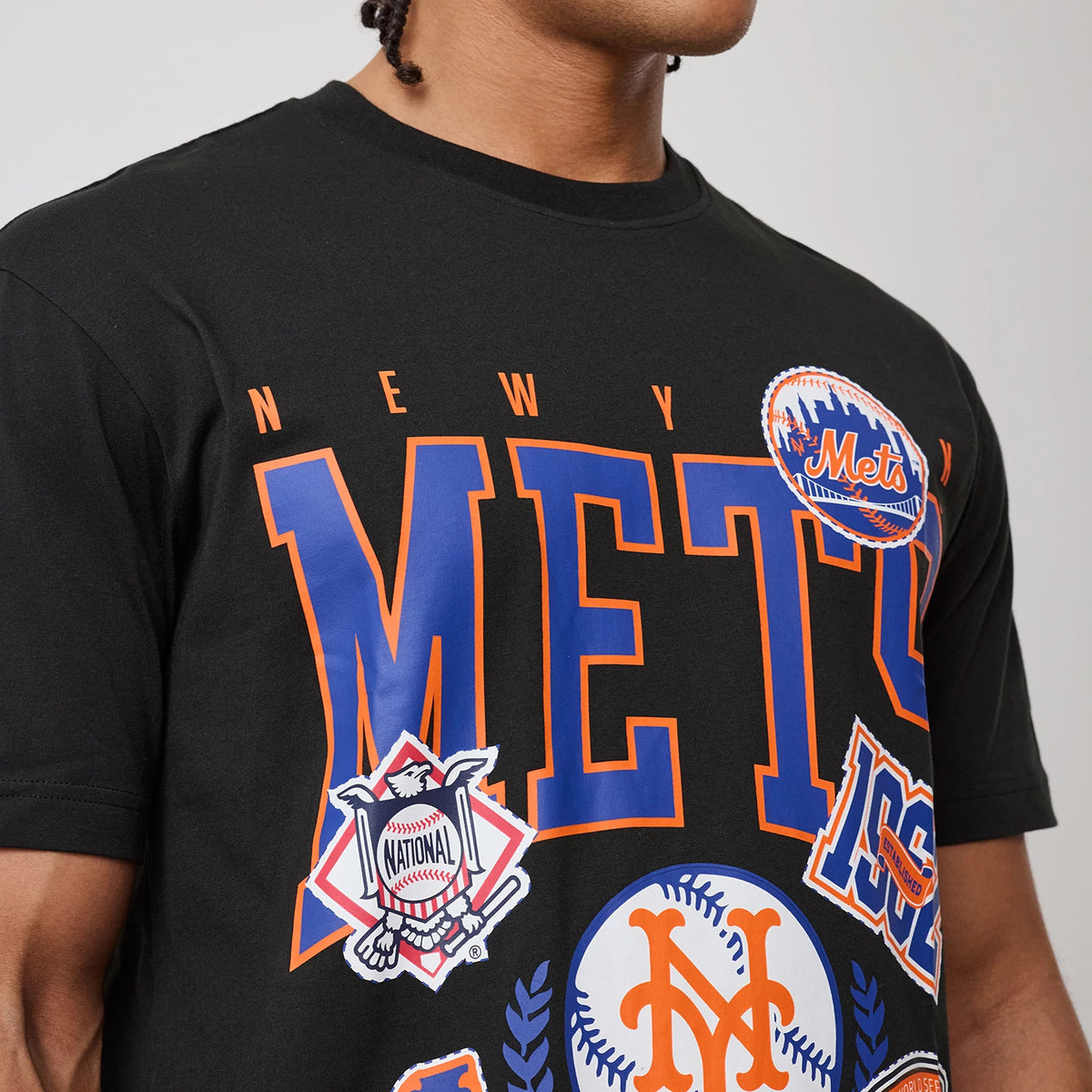 New Era New York Mets MLB Sport Classic Black T-Shirt, front view on-model showing large Mets lettering in blue and orange across the chest, with additional team graphics and a Mets logo badge on the upper right side.
