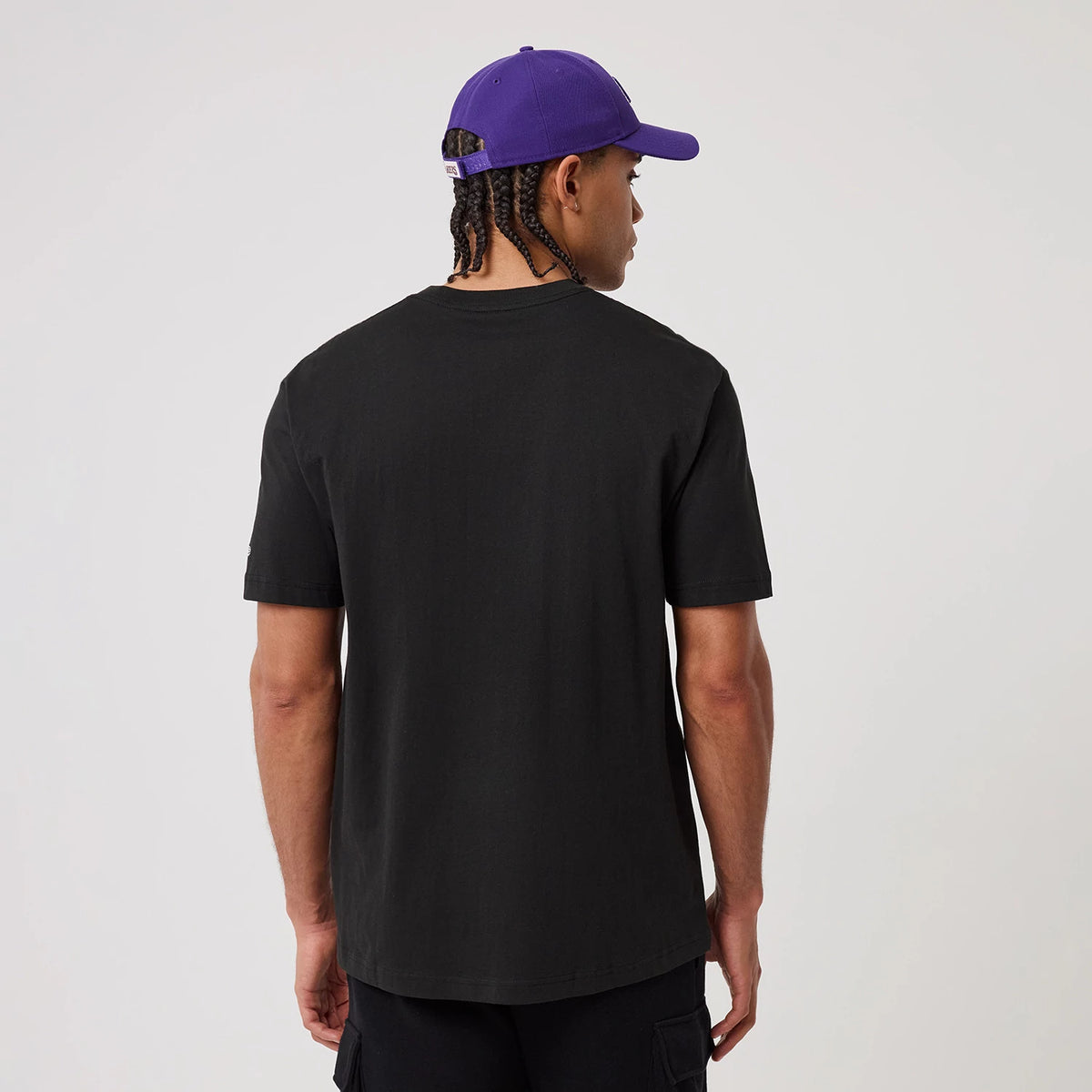 New Era LA Lakers NBA Sport Classic Black T-Shirt, back view on-model showing plain black fabric with short sleeves, styled with a purple cap displaying an embroidered logo on the back above adjustable strap.