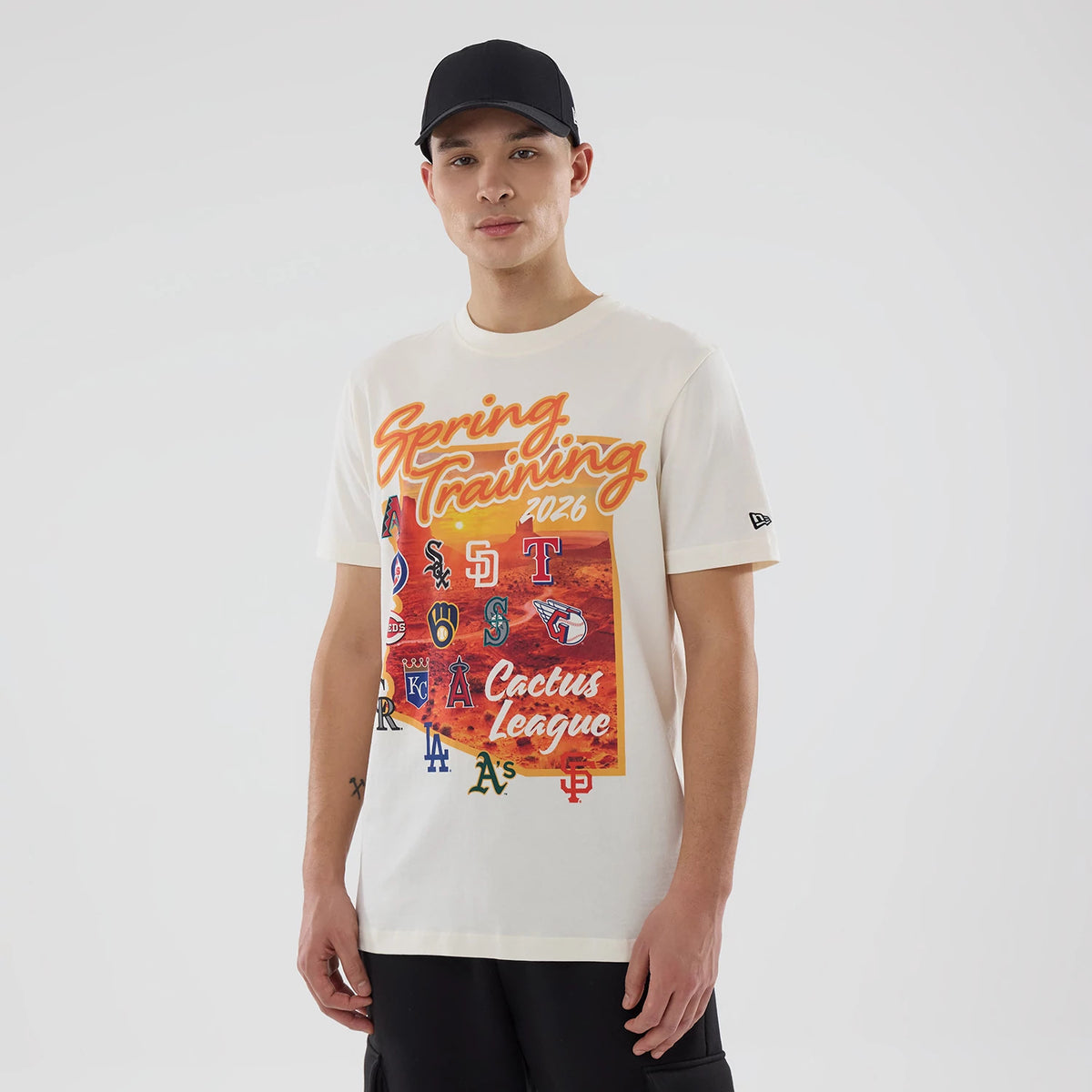 New Era Cactus League Logo MLB Spring Training 2026 Cream T-Shirt, front view on-model featuring 'Spring Training 2026' text above various MLB team logos in colours, New Era embroidered logo on the left sleeve.