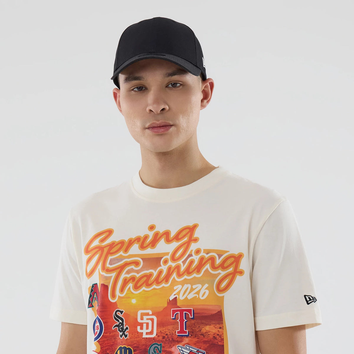 New Era Cactus League Logo MLB Spring Training 2026 Cream T-Shirt, front view on a model with a black cap, featuring Spring Training 2026 lettering in orange across the chest above a colourful graphic of MLB team logos.
