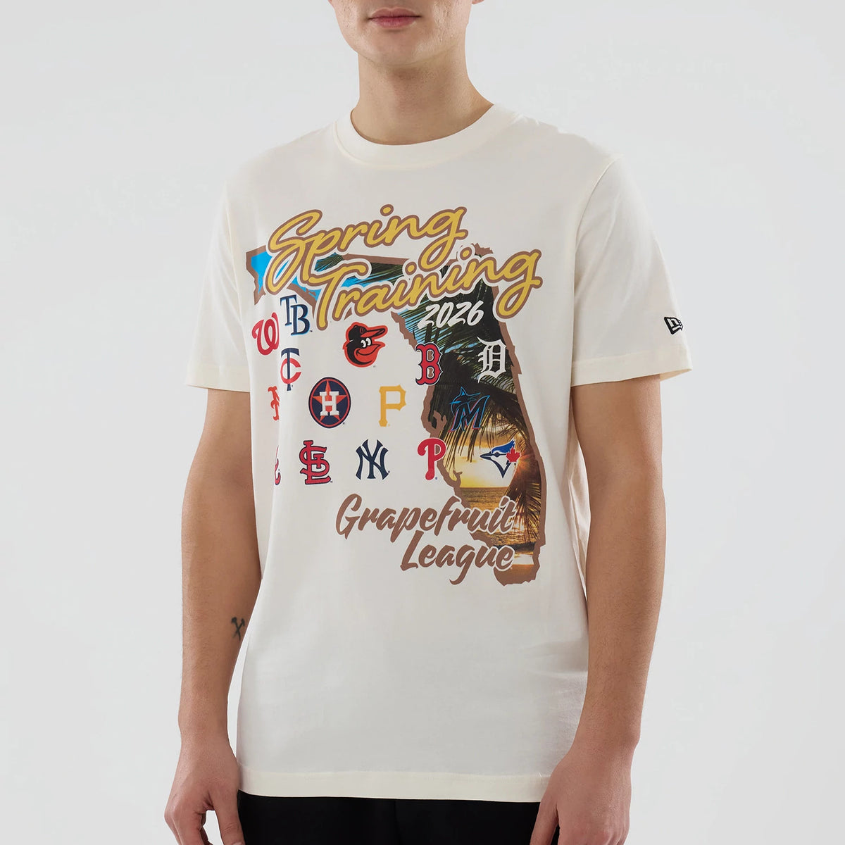 New Era Grapefruit League Logo MLB Spring Training 2026 Cream T-Shirt, front view on-model showing a colourful graphic with multiple MLB team logos, Spring Training 2026 text in yellow, and New Era embroidered logo on the left sleeve.