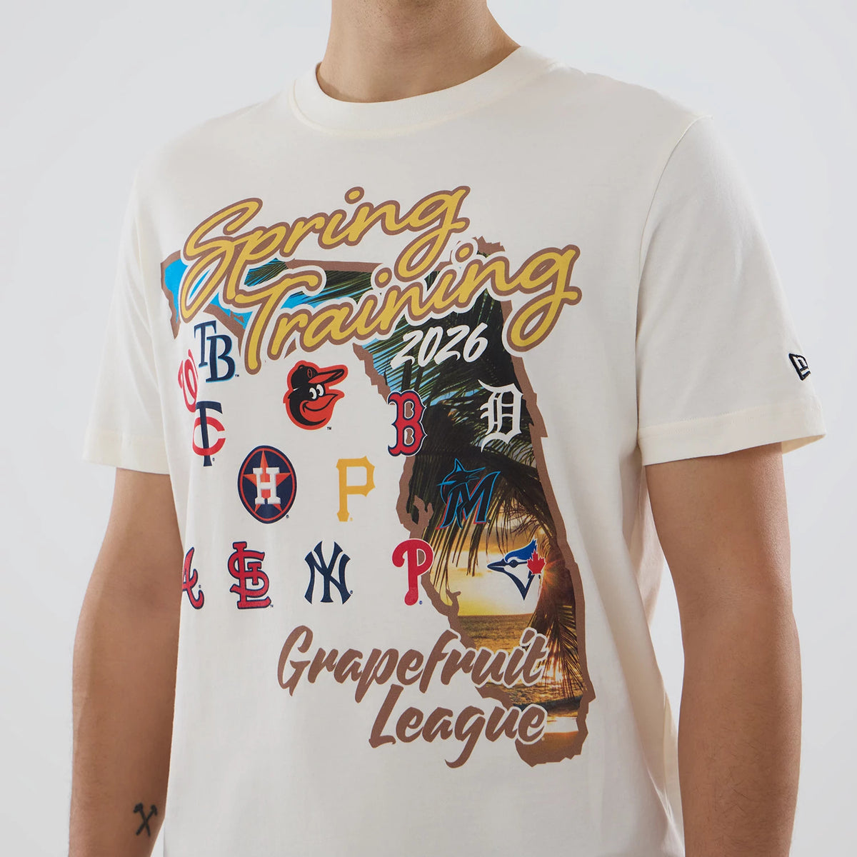 New Era Grapefruit League Logo MLB Spring Training 2026 Cream T-Shirt, front view showing graphics of the words Spring Training and Grapefruit League with various team logos, and an embroidered New Era logo on the sleeve.