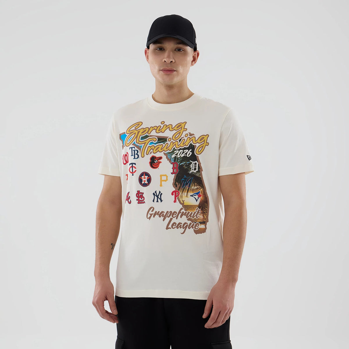 New Era Grapefruit League Logo MLB Spring Training 2026 Cream T-Shirt, front view on a model featuring multiple MLB team logos, large Spring Training lettering and a graphic of a player, with a black New Era cap.