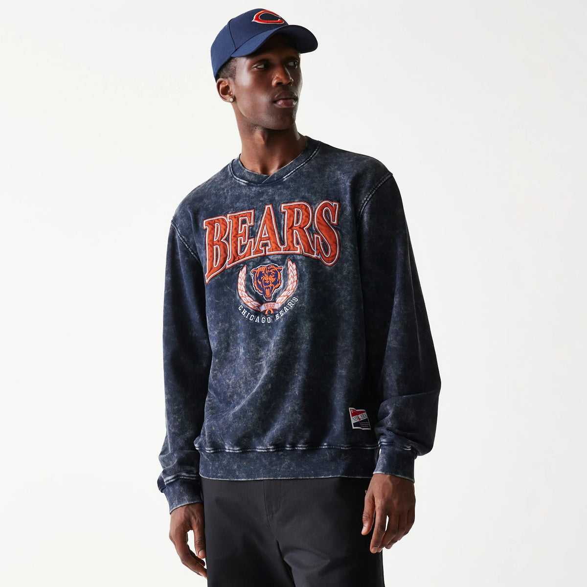 New Era Chicago Bears NFL Navy Crew Neck Sweater, front view on a model wearing a matching cap, featuring large orange Bears text with the team logo and a wreath design on the chest, rendered in distressed navy fabric.