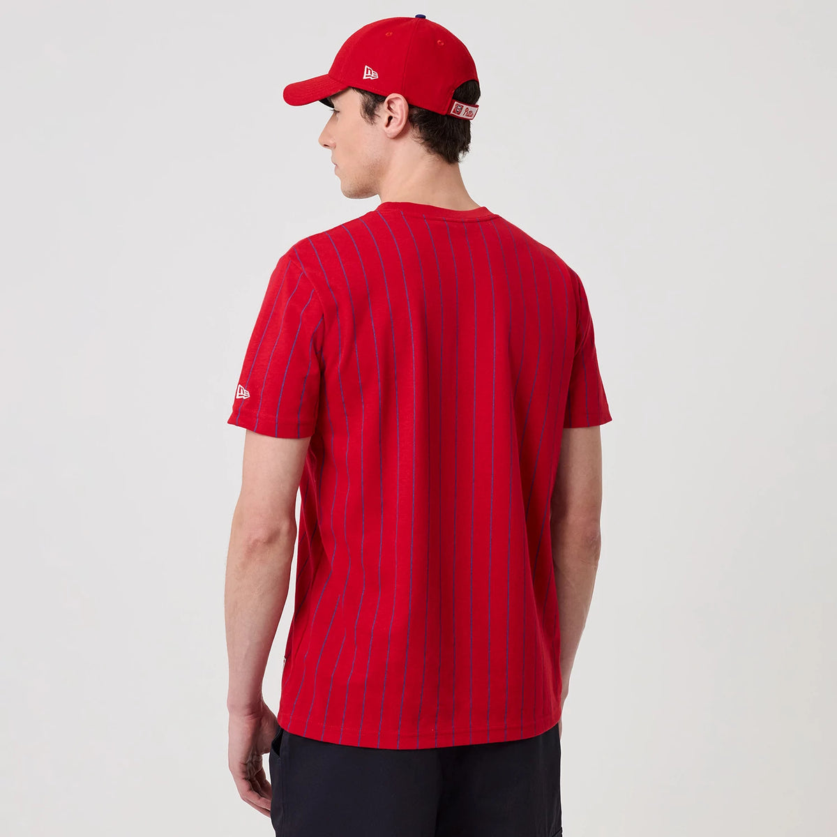 New Era Philadelphia Phillies MLB Pinstripe Red T-Shirt, back view on a model paired with a red cap, showing thin blue pinstripes running vertically down the bright red fabric and an embroidered New Era logo on the left sleeve.