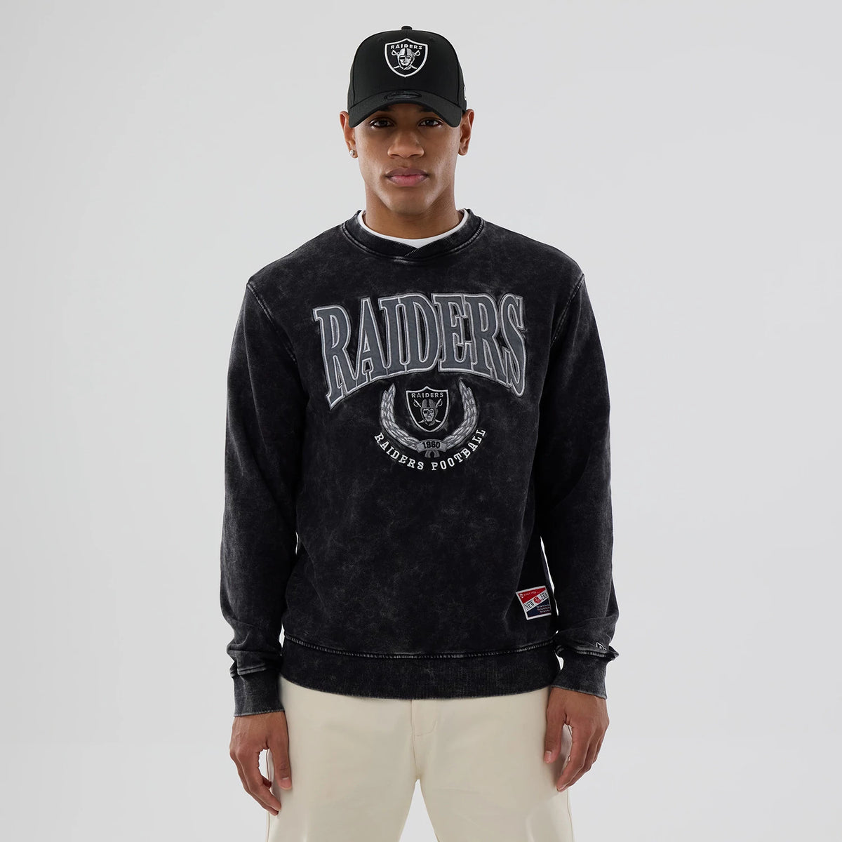 New Era Las Vegas Raiders NFL Laurel Wreath Black Crew Neck Sweater, front view on a model wearing a matching cap, featuring large grey Raiders lettering and emblem embroidery on the chest above a small team patch near the hem.