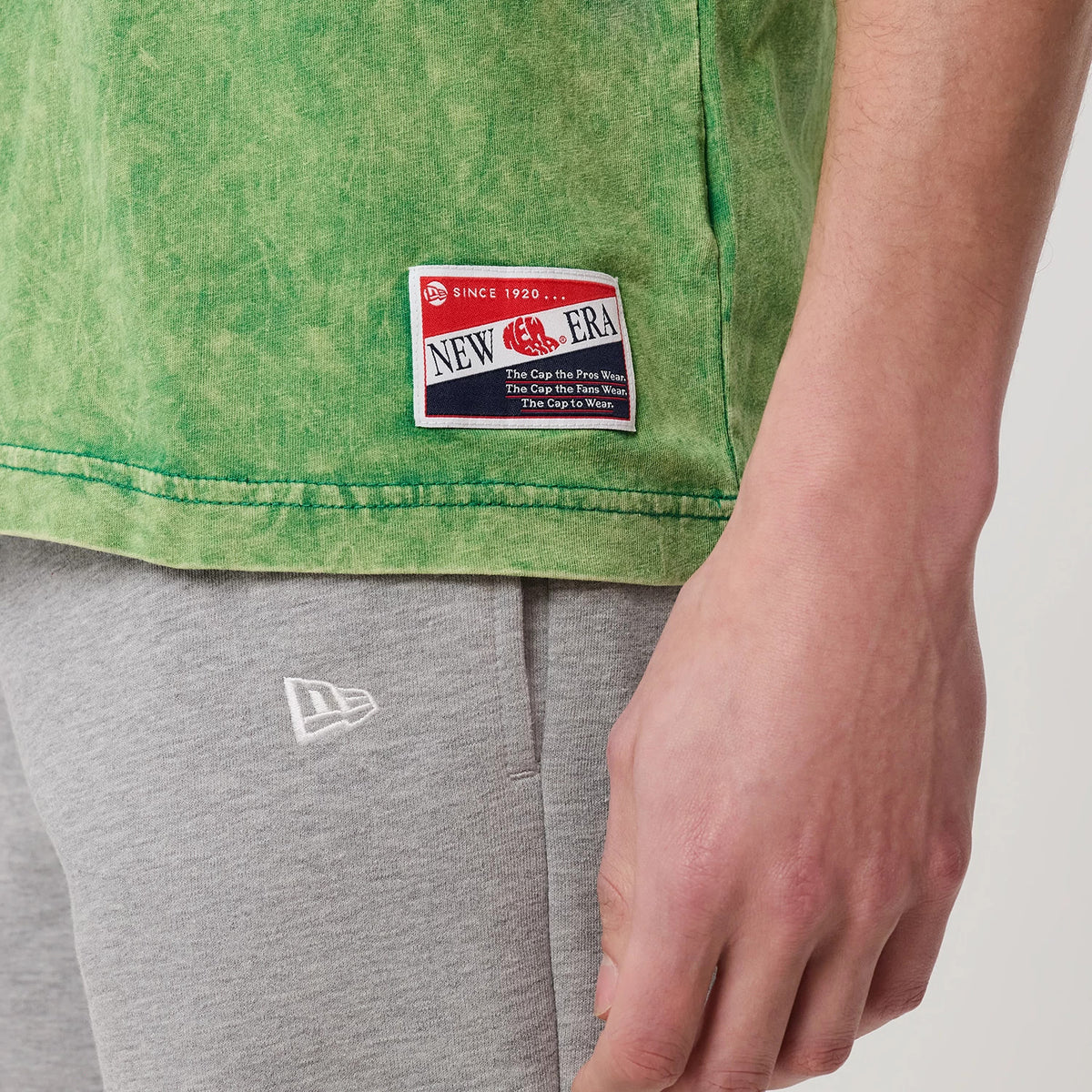 New Era Boston Celtics NBA Green T-Shirt, side view showing a large woven New Era logo patch near the hem on green fabric, styled with grey trousers featuring an embroidered white New Era logo on the pocket.