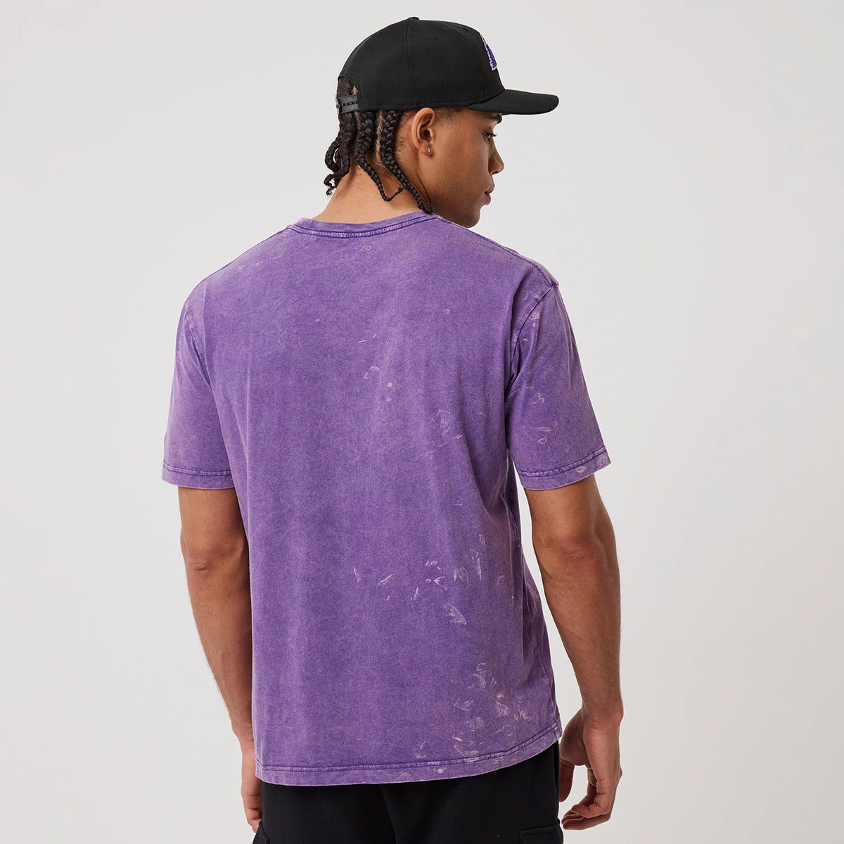 New Era LA Lakers NBA Purple T-Shirt, back view depicting a plain purple tee with a slightly faded effect, worn by a model paired with black shorts and a black cap for a casual look.