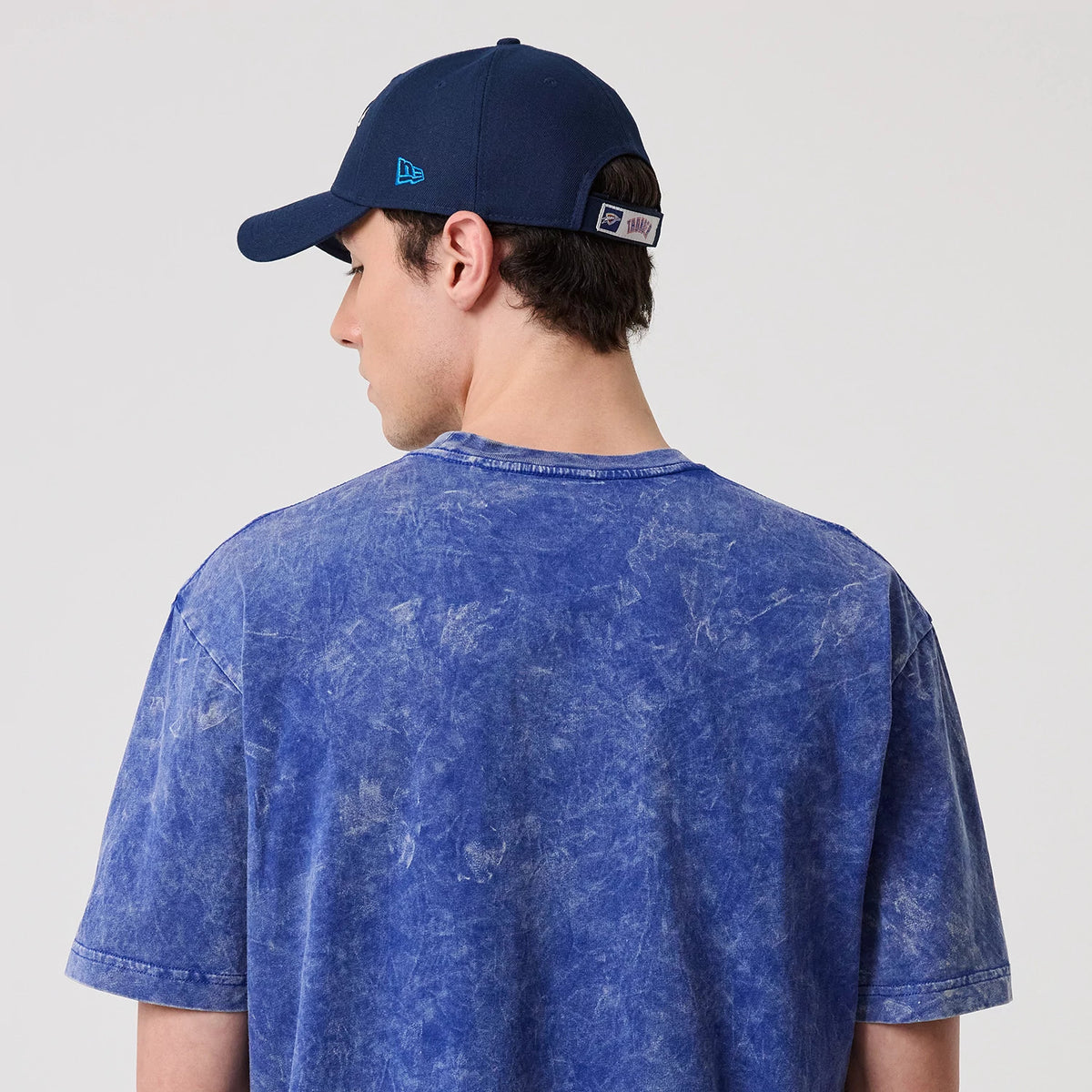 New Era Oklahoma City Thunder NBA Blue T-Shirt, back view on a model, showing a marbled blue design and relaxed fit with the model wearing a matching dark blue cap featuring an embroidered New Era logo on the side.