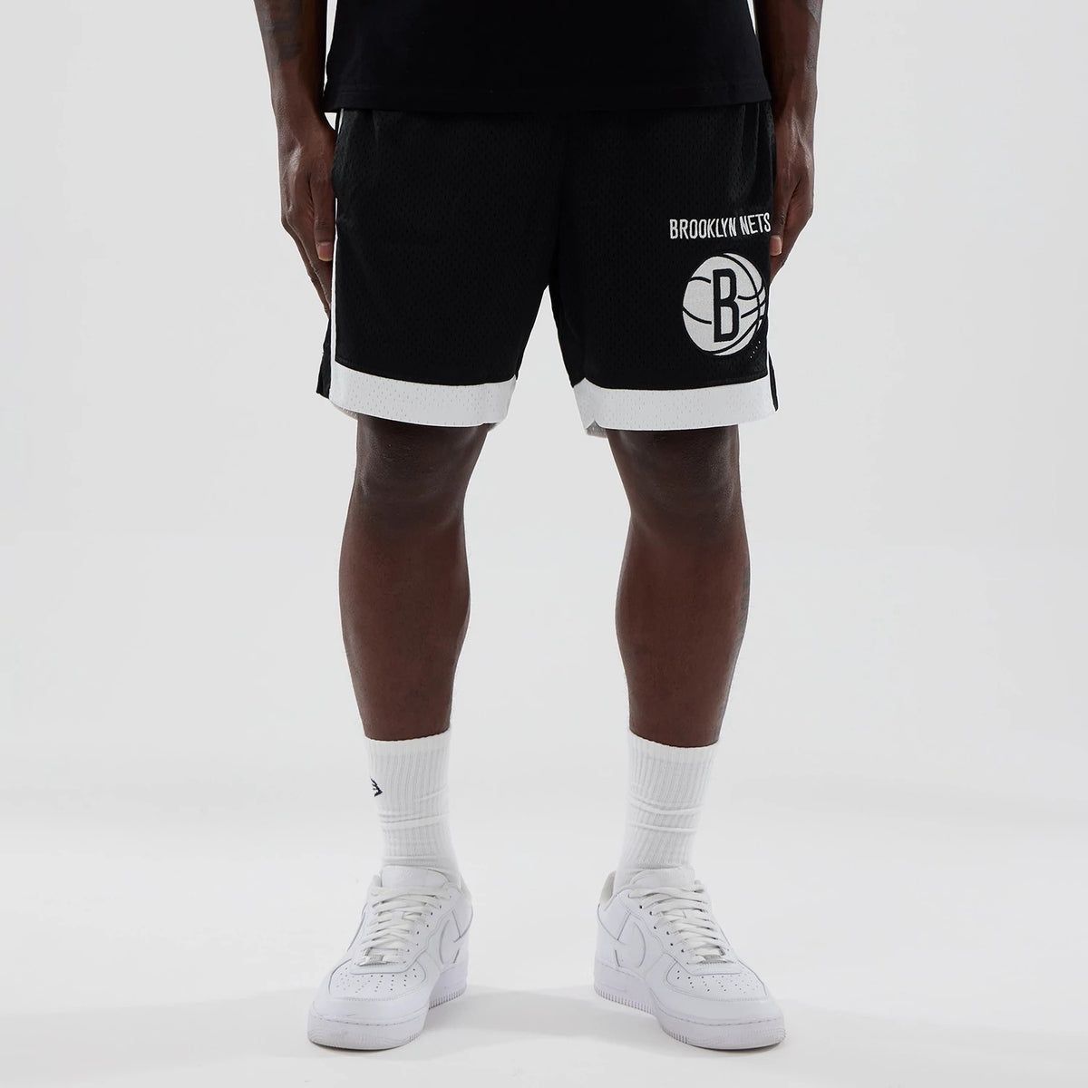 New Era Brooklyn Nets NBA Black Mesh Shorts, front view on a model showing the white embroidered Brooklyn Nets logo on the lower left side, with white trim at the hem and solid black mesh fabric for the main body.