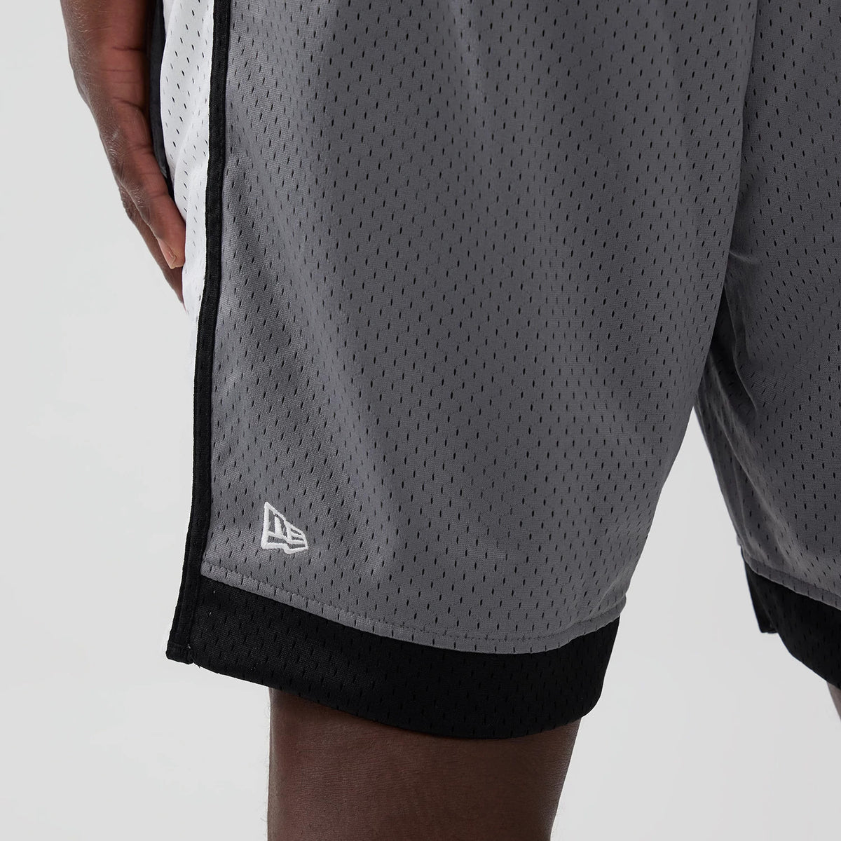New Era Las Vegas Raiders NFL Grey Mesh Shorts, side view showing the light grey mesh material with small perforations, a white embroidered New Era logo near the hem, and contrasting black trim along the bottom edge.