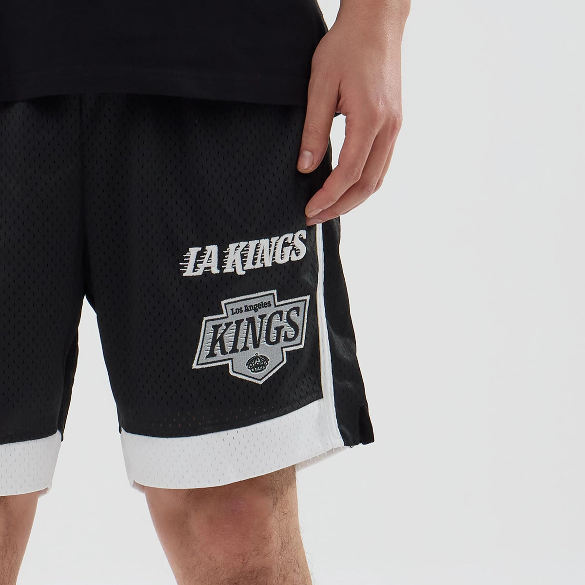 New Era LA Kings NHL Black Mesh Shorts, front view showing the black mesh fabric with white embroidered LA Kings lettering and a large Kings logo on the left leg, accented by white trim at the hem and side.