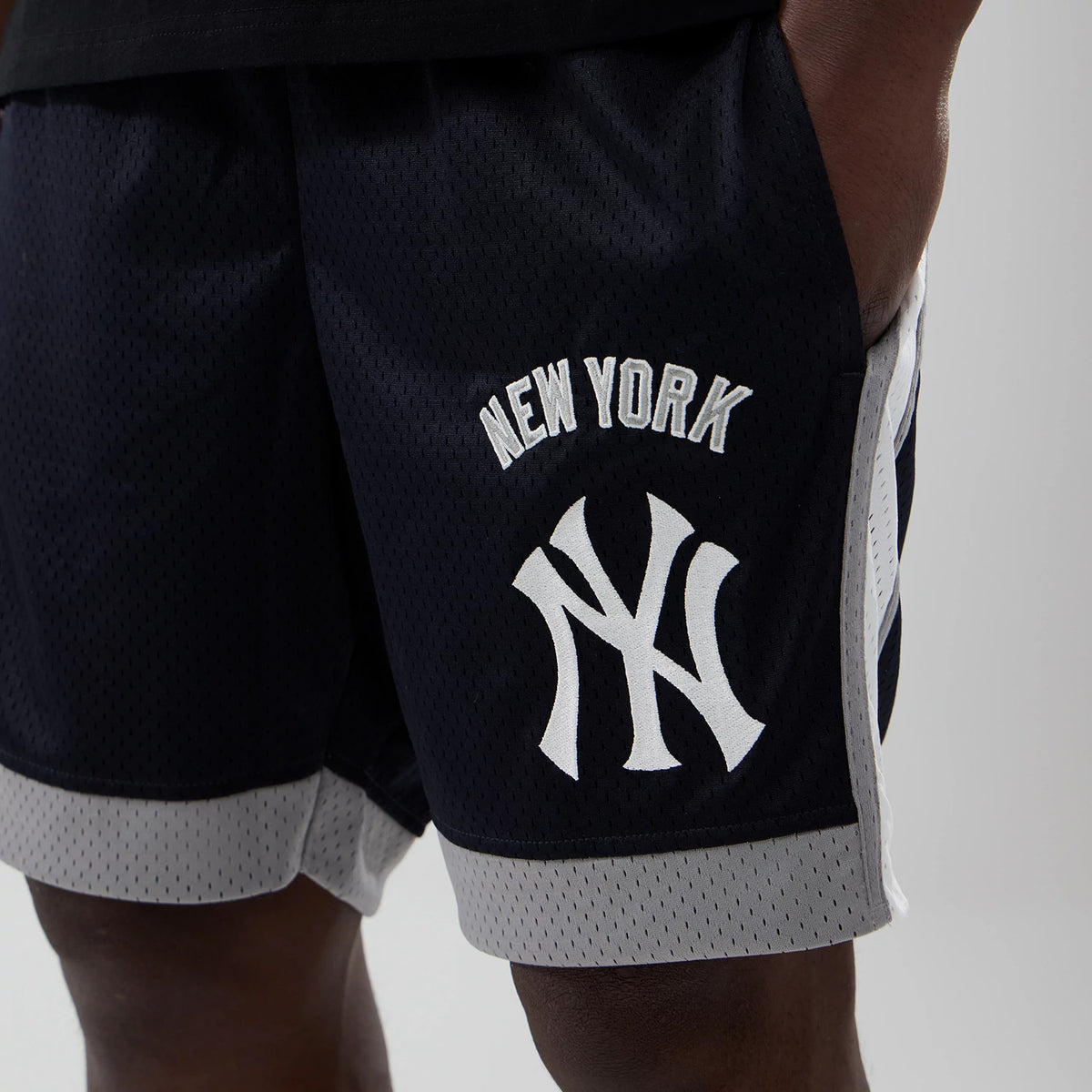 New Era New York Yankees MLB Navy Mesh Shorts, front view showing white embroidered New York lettering and Yankees logo on the navy fabric, grey trim along the hem and sides, styled casually on a model with hands in pockets.