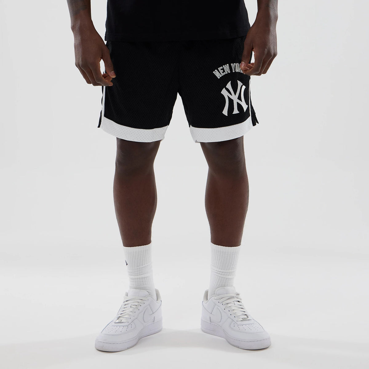 New Era New York Yankees MLB Black Mesh Shorts, front view on a model with white trainers and socks, featuring a large white embroidered Yankees logo on the left leg and contrasting white trim on the hem.
