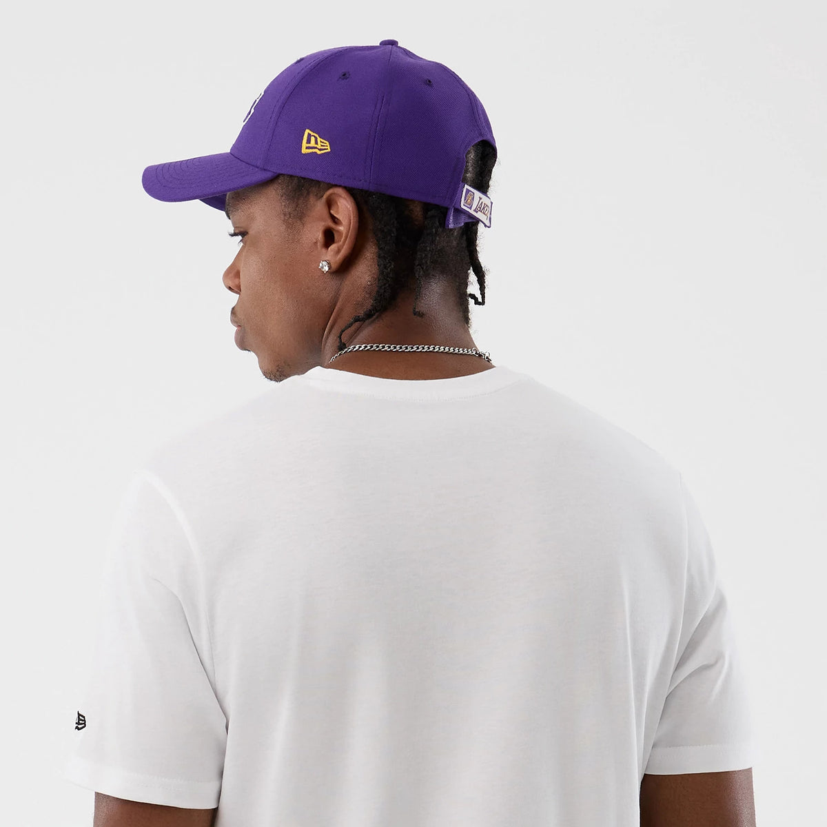 New Era LA Lakers NBA Script Back To School White T-Shirt, back view on-model showing a plain white back with a small embroidered New Era logo on the left sleeve, and styled with a purple cap featuring a yellow embroidered New Era logo on the side.