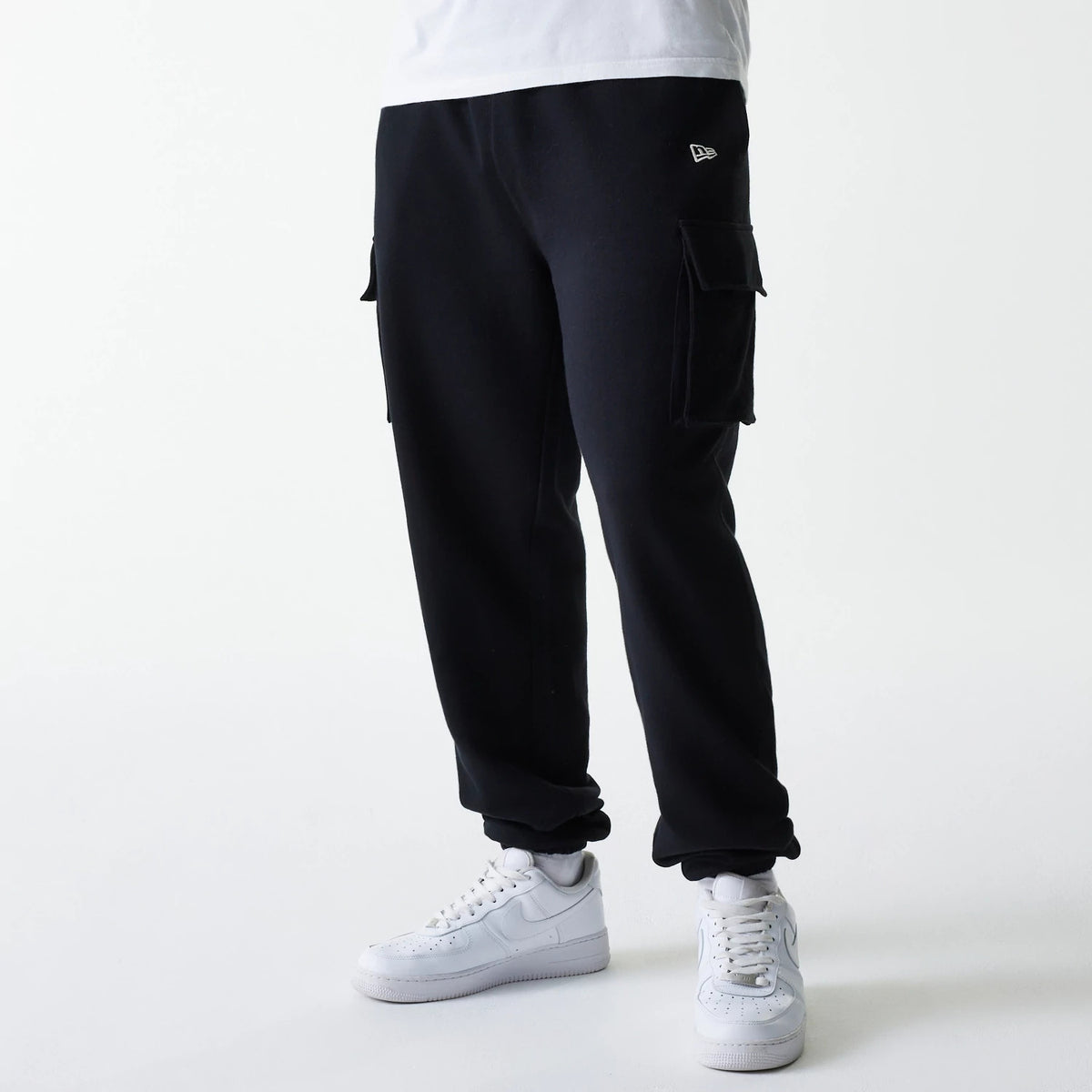 New Era Black Cargo Joggers, front view on a model wearing white trainers and a white T-shirt, showing the embroidered New Era flag logo on the left upper thigh and two large flap pockets on the sides of the black joggers.