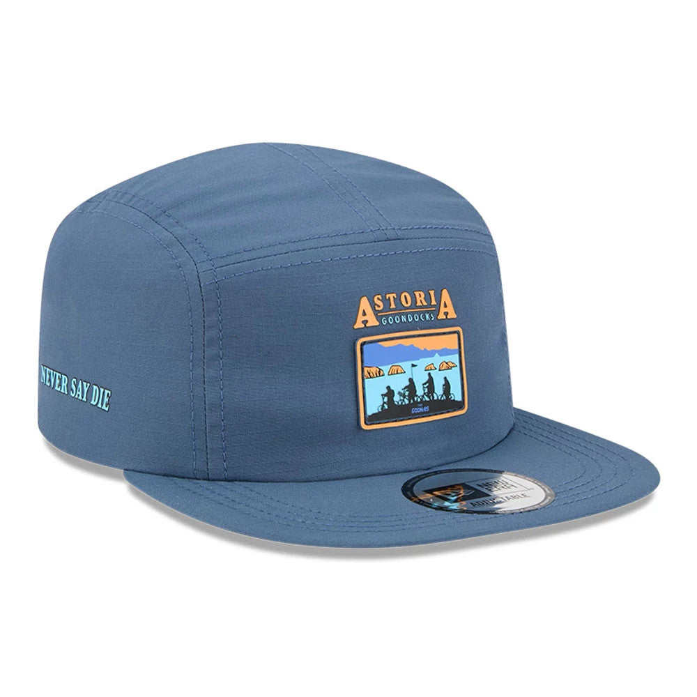 New Era The Goonies Astoria Warner Brothers Open Blue Camper Cap, three-quarter view showing an embroidered Astoria logo patch on the centre front panel, flat blue visor with a 9FORTY size sticker, and Never Say Die text on the left side.