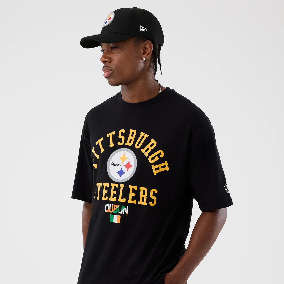 New Era Pittsburgh Steelers NFL International Series Games Black Oversized T-Shirt, front view on a model wearing a black cap, featuring Pittsburgh Steelers lettering in yellow with a team logo at the centre and Dublin text with an Irish flag below.