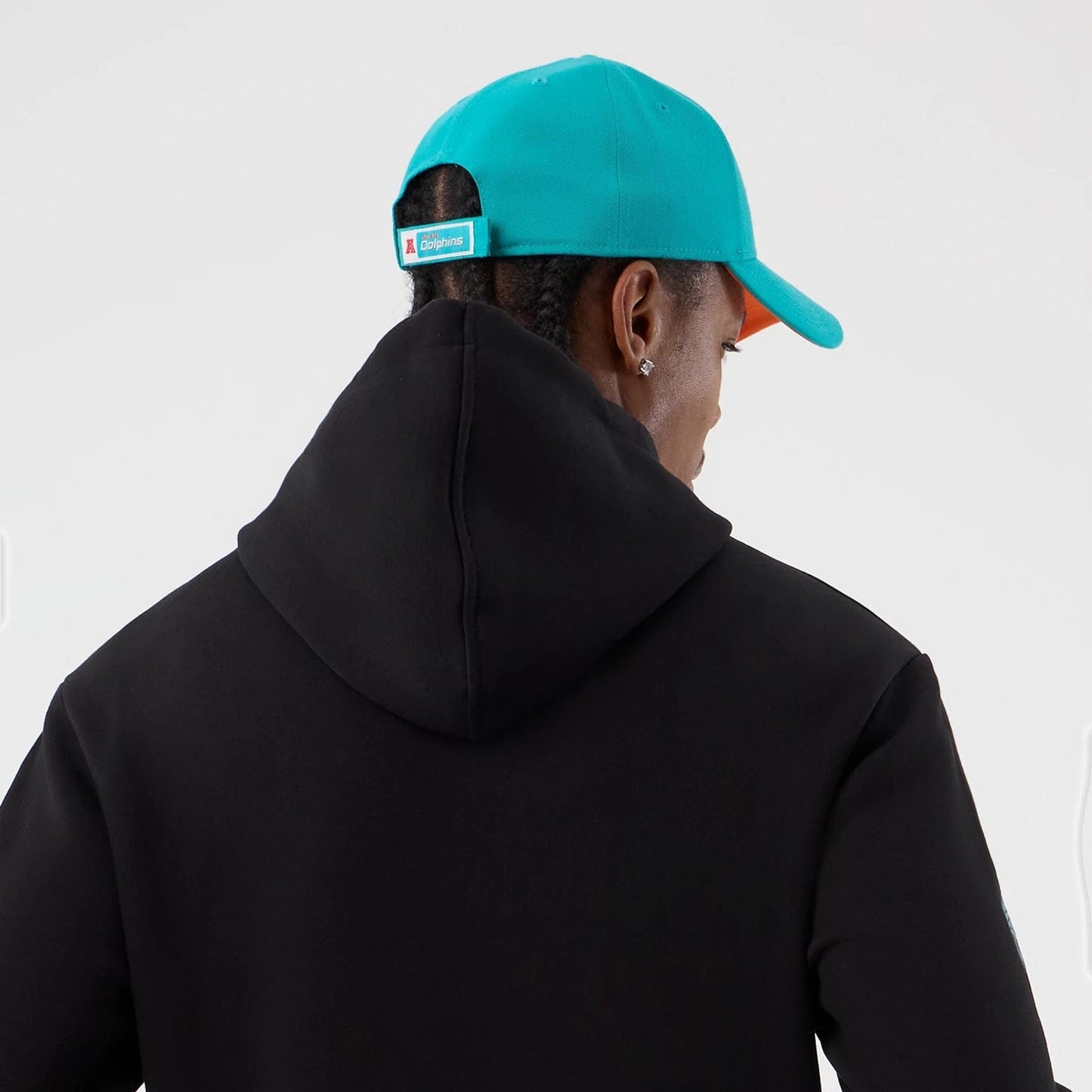 New Era Dolphins NFL International Series Games Patch Black Pullover Hoodie, back view on-model, showing the black hood draped across the shoulders with the model wearing a bright turquoise cap featuring a Dolphins label above the closure.