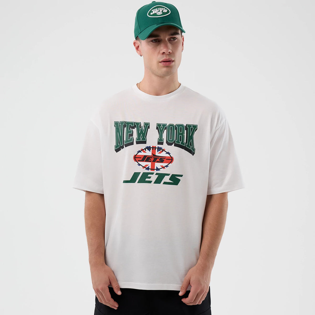 New Era New York Jets NFL International Series Games White Oversized T-Shirt, front view on-model highlighting the large New York Jets lettering centred on the chest in green above a stylised globe graphic and Jets text, paired with a matching cap.