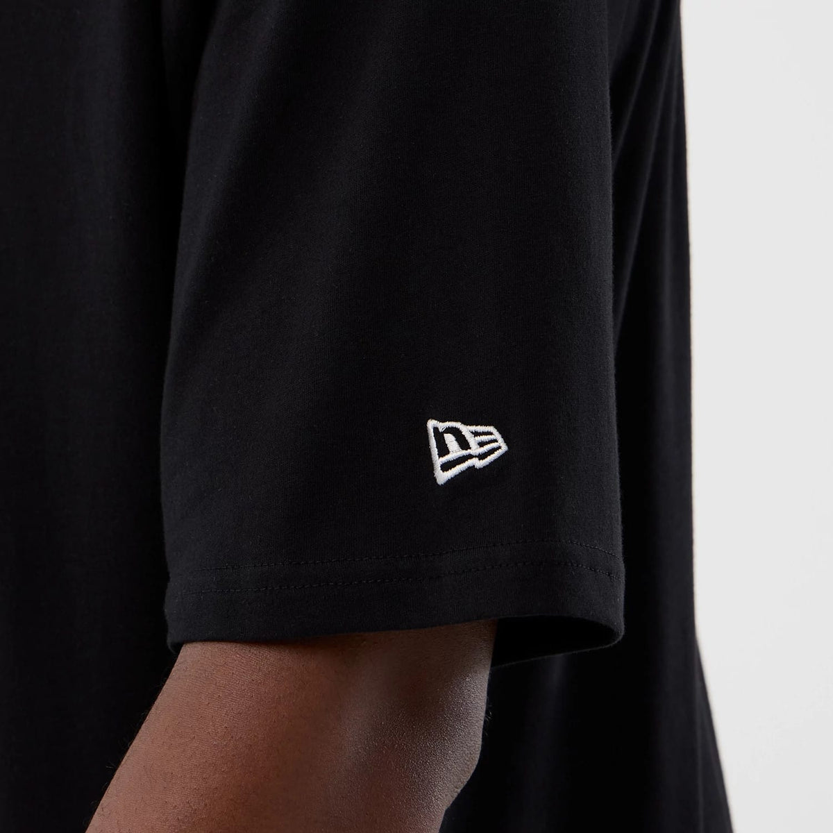 New Era Berlin NFL International Series Games Black Oversized T-Shirt, side view showing the sleeve with a white embroidered New Era logo near the edge against the plain black fabric.