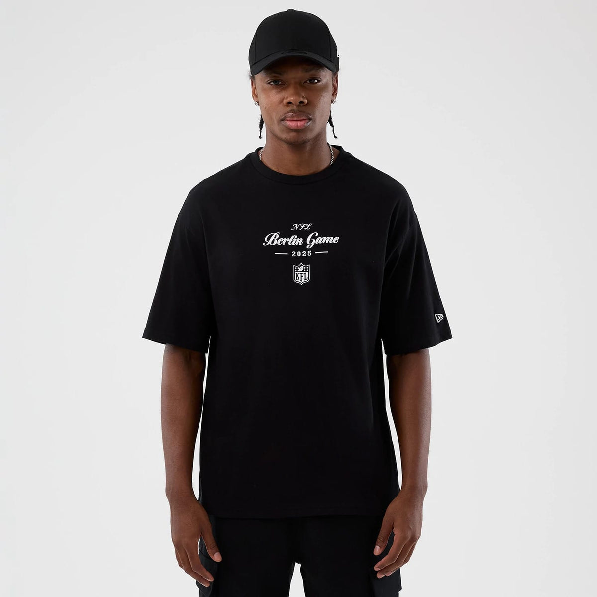 New Era Berlin NFL International Series Games Black Oversized T-Shirt, front view on a model paired with a plain black cap, featuring a central white printed Berlin Game 2023 graphic and a small embroidered New Era logo on the left sleeve.
