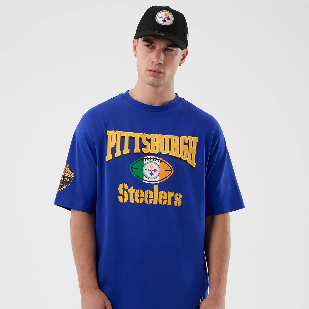 New Era Pittsburgh Steelers NFL International Series Games Blue Oversized T-Shirt, front view on a model wearing a matching cap, featuring a large yellow Pittsburgh Steelers graphic across the chest and a team logo on the left sleeve.