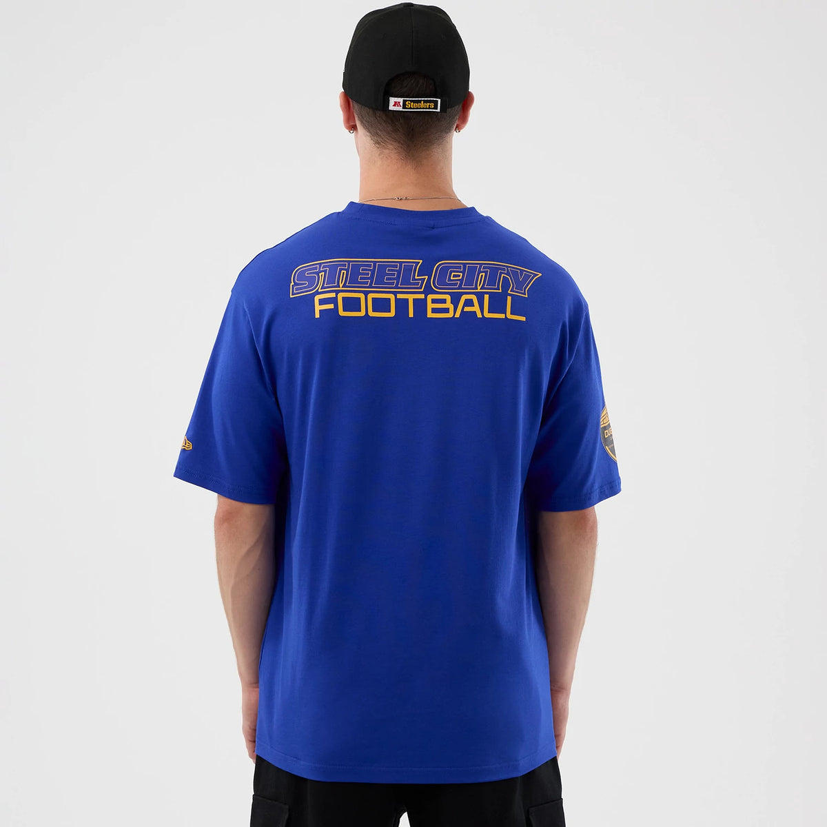 New Era Pittsburgh Steelers NFL International Series Games Blue Oversized T-Shirt, back view on-model showing Steel City Football lettering across the shoulders in purple and yellow, and a patch on the left sleeve.