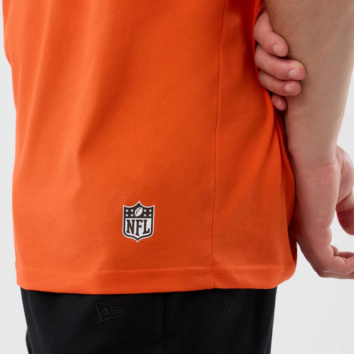 New Era Cleveland Browns NFL International Series Games Orange Oversized T-Shirt, side view showing the orange fabric with a printed NFL logo near the hem and a New Era logo near the bottom edge.