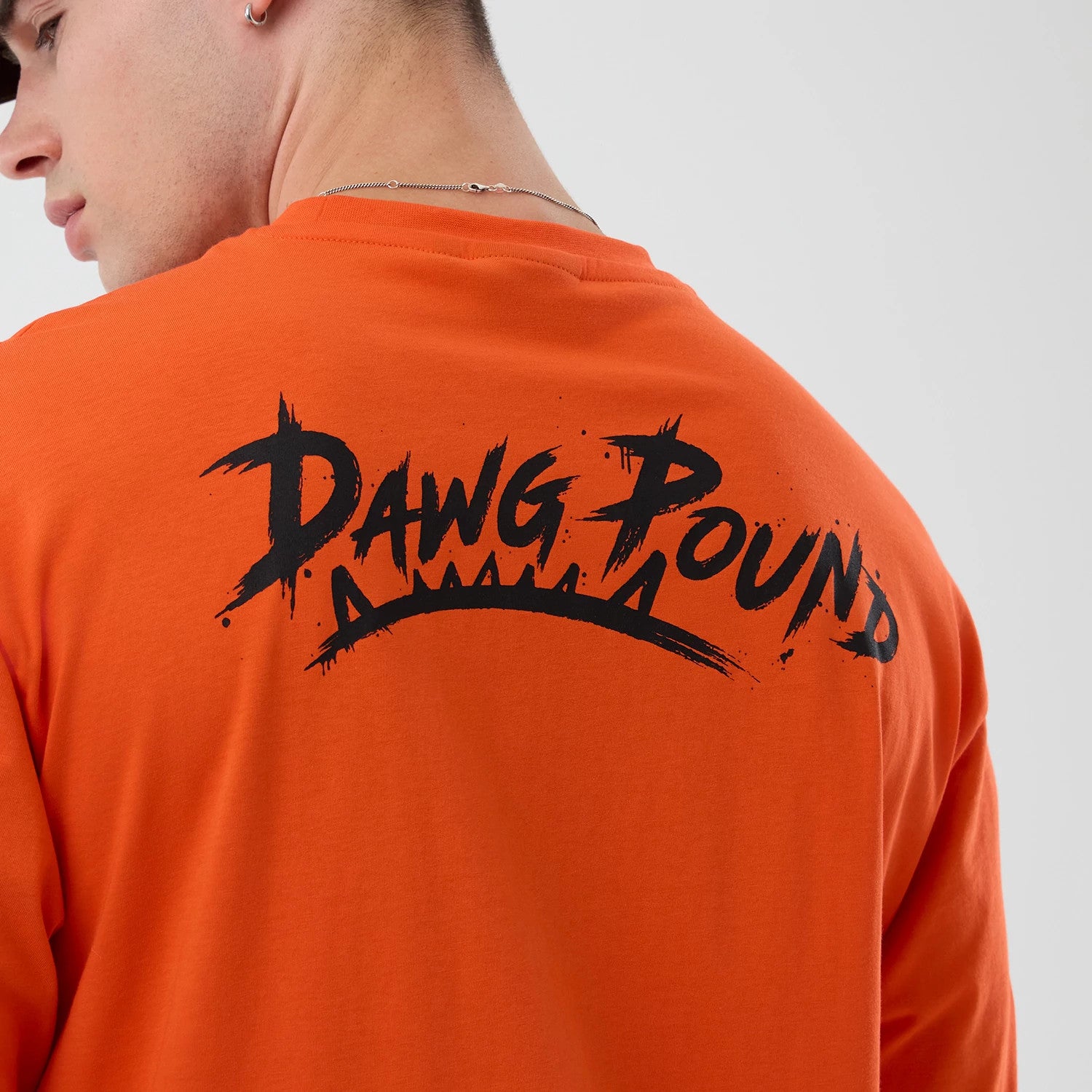 New Era Cleveland Browns NFL International Series Games Orange Oversized T-Shirt, back view on a model showing black Dawg Pound lettering with graphic spikes underneath, set against the bright orange fabric.
