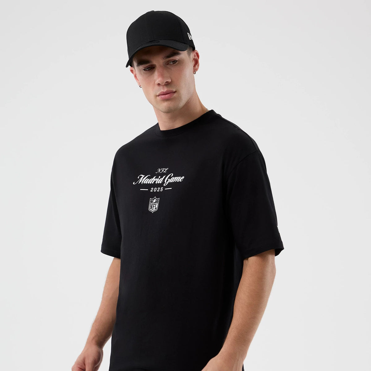 New Era Madrid Game NFL International Series Games Black Oversized T-Shirt, front view on a model with a black cap, showing white printed Madrid Game 2025 lettering and NFL shield logo prominently on the chest.