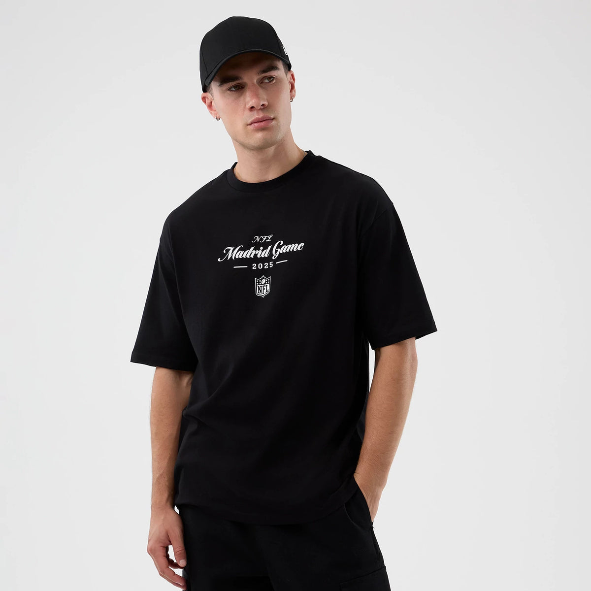New Era Madrid Game NFL International Series Games Black Oversized T-Shirt, front view on a model wearing a plain black cap, showcasing the white embroidered NFL Madrid Game 2025 lettering and the NFL shield graphic below on the chest.
