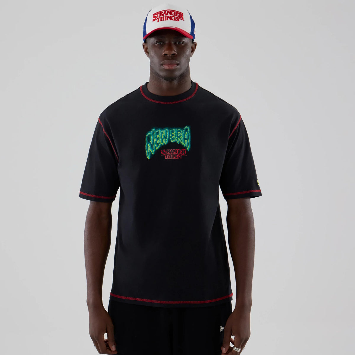 New Era Stranger Things Black Oversized T-Shirt, front view on-model showing a green embroidered New Era logo and red Stranger Things lettering on the chest, with red stitching along the seams and sleeves paired with a matching cap.